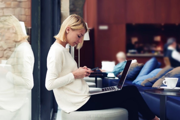 Woman sitting in a lobby, looking at her phone, and a laptop on her lap.