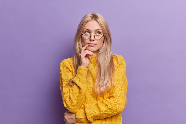 Young pretty young woman thinks of ideas concentrated above stands thoughtful and keeps hand on face stands in thoughtful pose wears round glasses yellow sweater isolated over purple background.