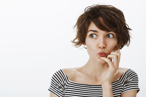 Indoor shot of curious emotive young female student with cute messy hairstyle, folding lips and saying wow while holding hand near mouth, looking left with interest and curiosity over grey wall.