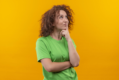 Young woman with short curly hair in green t-shirt looking with pensive expression on face thinking standing over orange wall